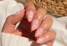 woman’s hand featuring cute summer July nail design with glossy coral French tips, white daisy flower nail art, almond-shaped gel nails, gold jewelry, and bright sunny natural lighting.