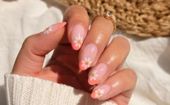 woman’s hand featuring cute summer July nail design with glossy coral French tips, white daisy flower nail art, almond-shaped gel nails, gold jewelry, and bright sunny natural lighting.