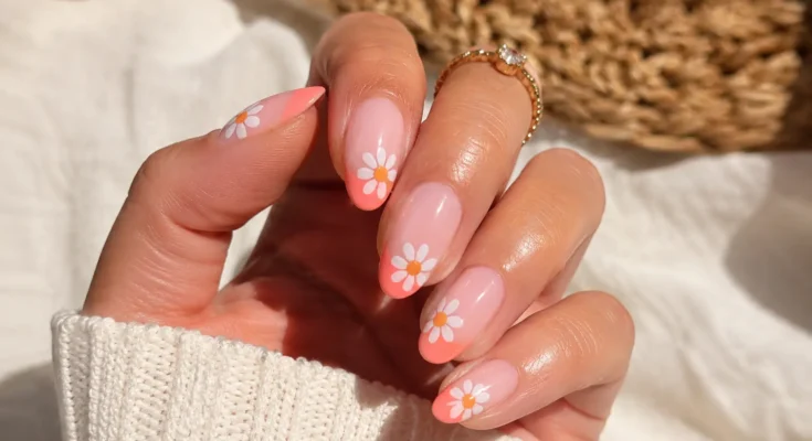 woman’s hand featuring cute summer July nail design with glossy coral French tips, white daisy flower nail art, almond-shaped gel nails, gold jewelry, and bright sunny natural lighting.