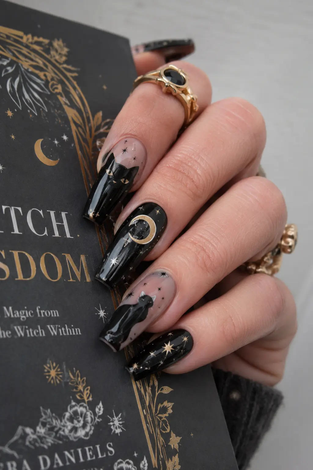 Black cat coffin nails with glossy moon and star designs, elegant rings, and a spooky manicure style while holding a book.