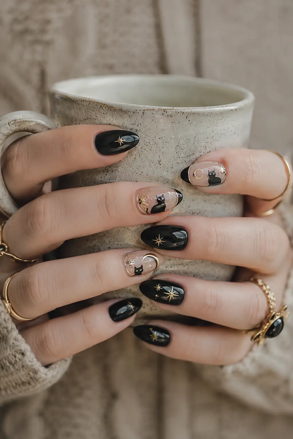 Close-up of glossy black cat nails with gold moon and star accents, gold rings, and both hands holding a ceramic cup.