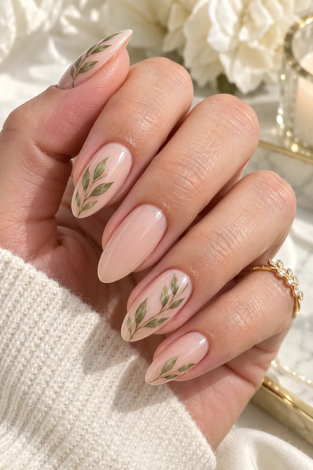 woman's hand with almond-shaped nude nails featuring delicate hand-painted olive green botanical leaf accents, photographed in bright natural daylight against a soft cream luxury background.