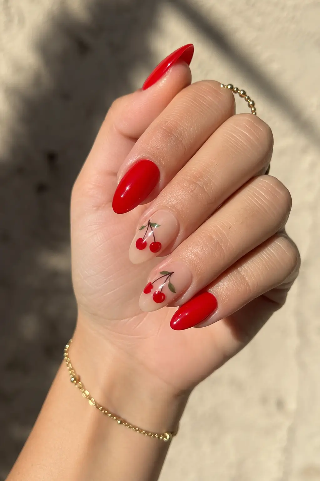 woman’s hand featuring glossy cherry red summer gel nails with tiny cherry nail art, almond-shaped manicure, smooth cuticles, delicate gold jewelry, and natural sunlight on a soft beige background.