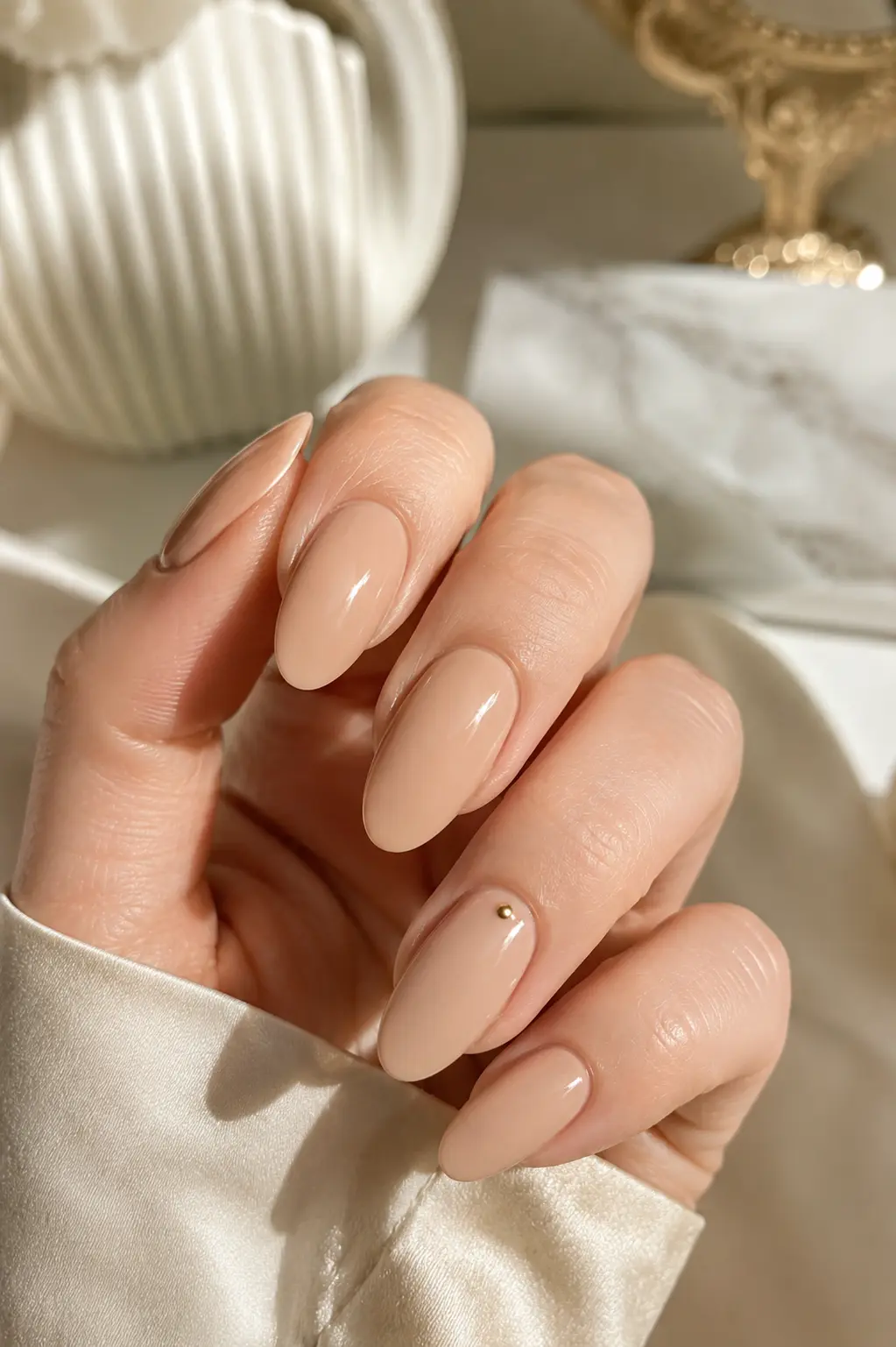 woman's hand with classic glossy nude almond-shaped nails featuring a tiny gold accent near one cuticle, captured in bright natural daylight against an elegant cream and ivory luxury background.