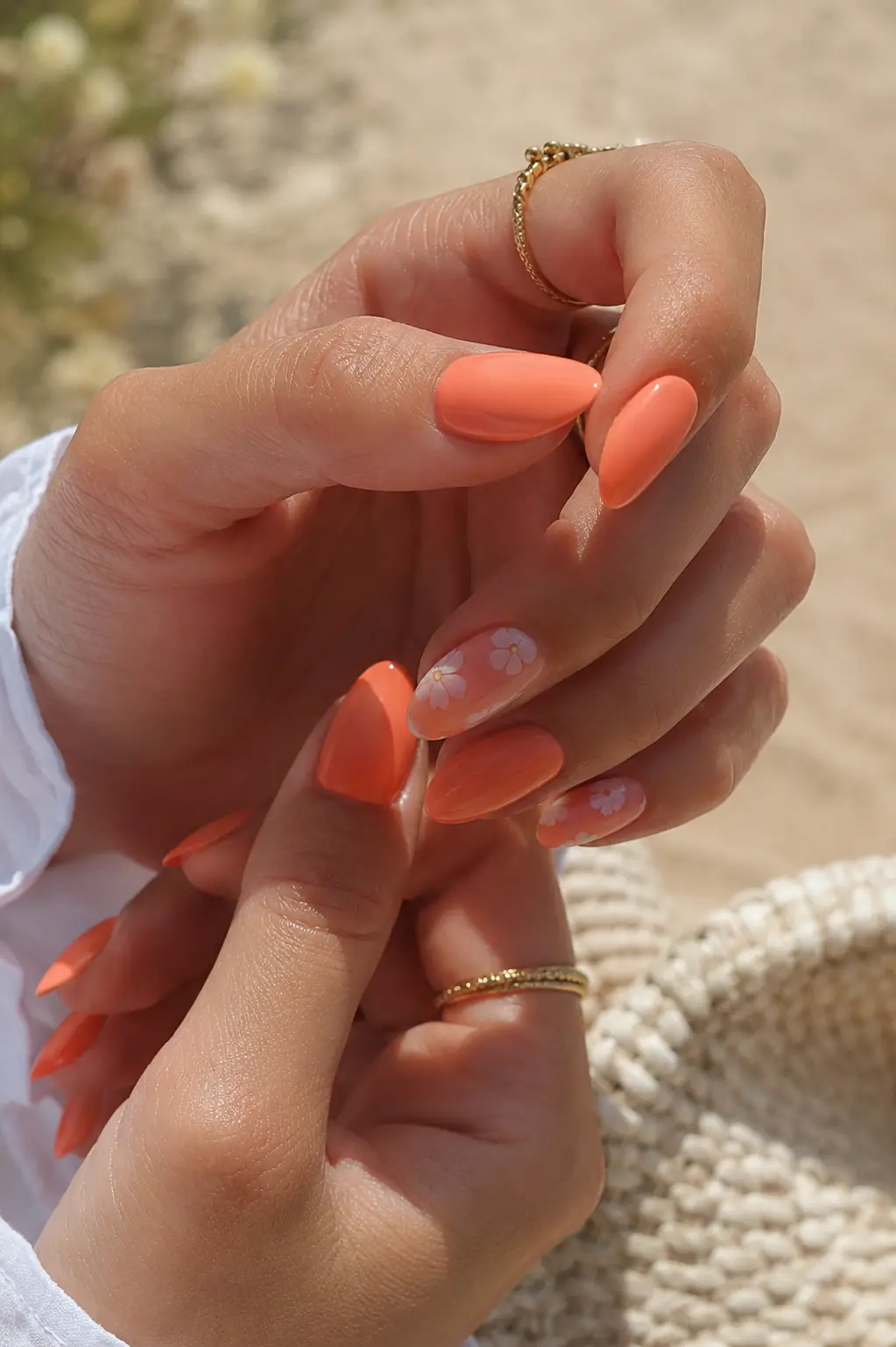 woman’s hands with glossy coral orange almond nails, subtle white floral nail art, smooth cuticles, gold rings, and natural skin texture in bright summer sunlight with a soft beige background.