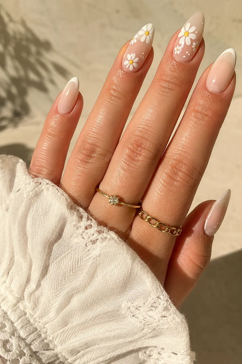 close-up of English woman’s hands with nude pastel almond nails, white daisy flower nail art, yellow centers, glossy gel finish, gold rings, and soft summer sunlight.