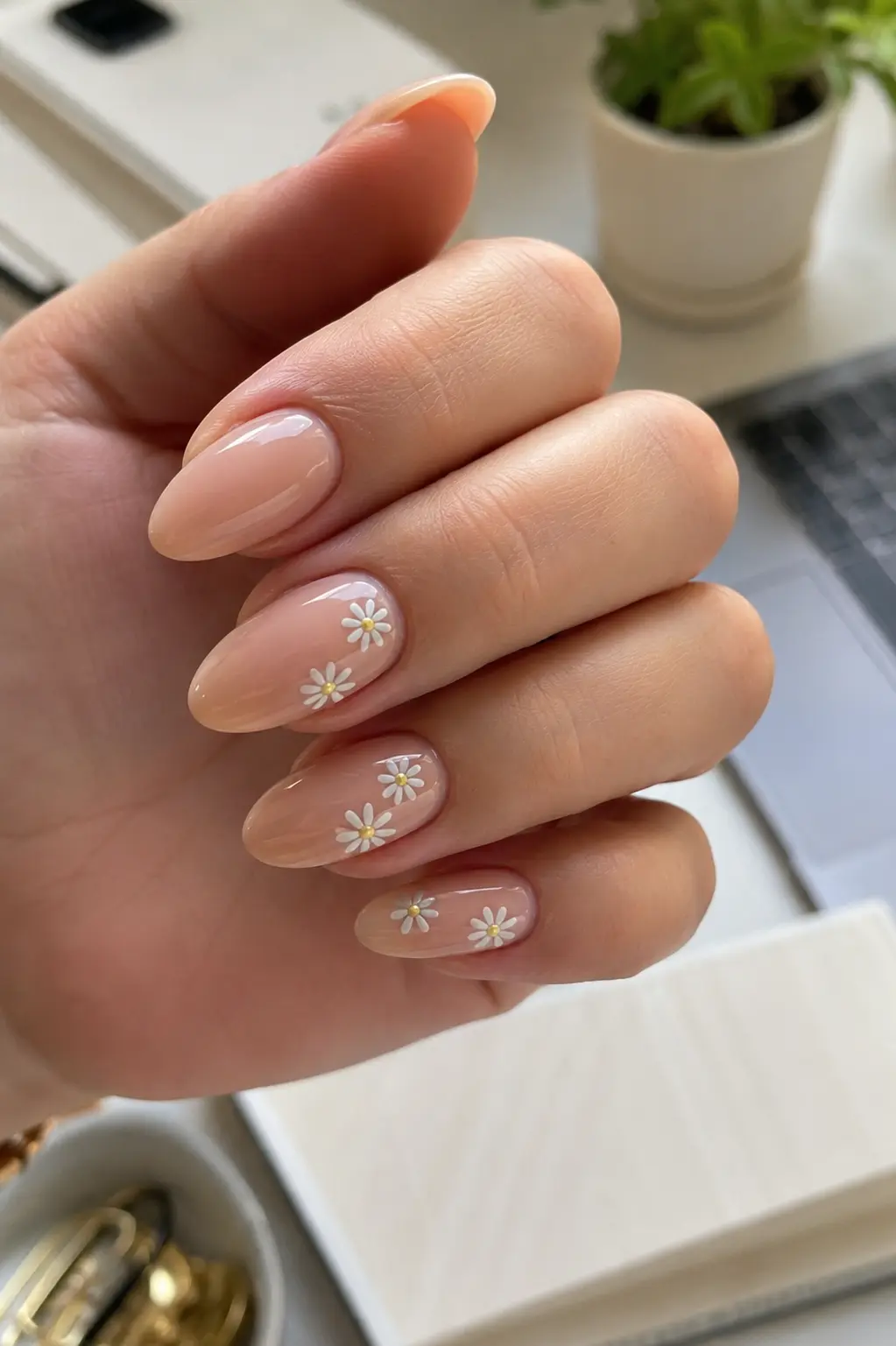 woman's hand showcasing glossy nude almond-shaped nails with delicate hand-painted white daisy accents.