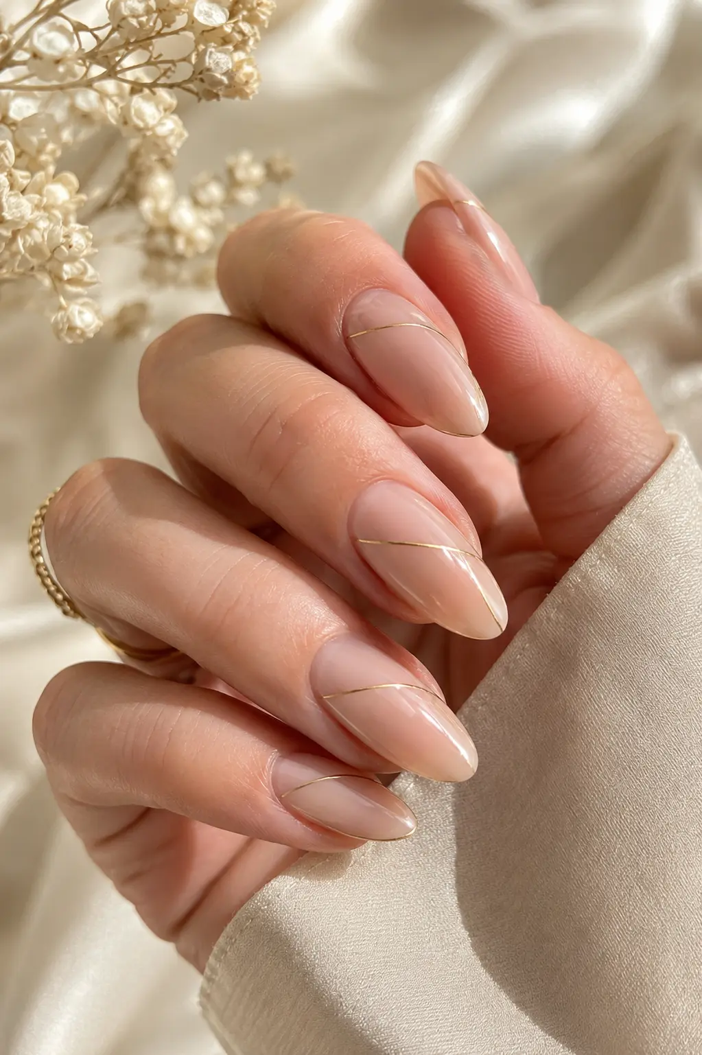 woman's hand with glossy nude almond-shaped nails featuring ultra-thin metallic gold line accents, photographed in bright natural daylight against a luxurious cream background
