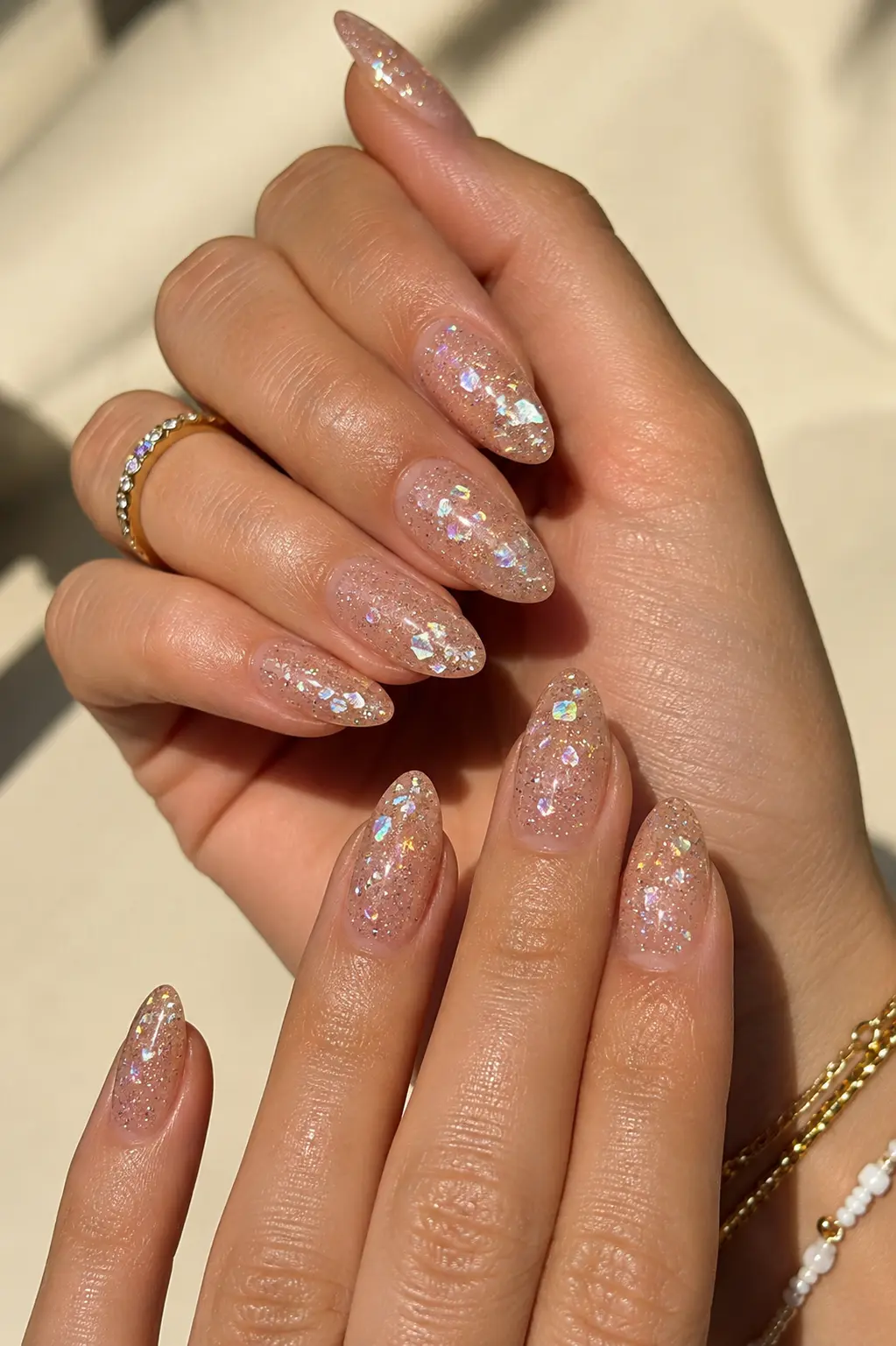 women’s hands featuring glitter sparkle summer July nail designs with glossy shimmer gel polish, almond-shaped nails, clean cuticles, gold jewelry, and bright natural sunlight on a soft beige background.