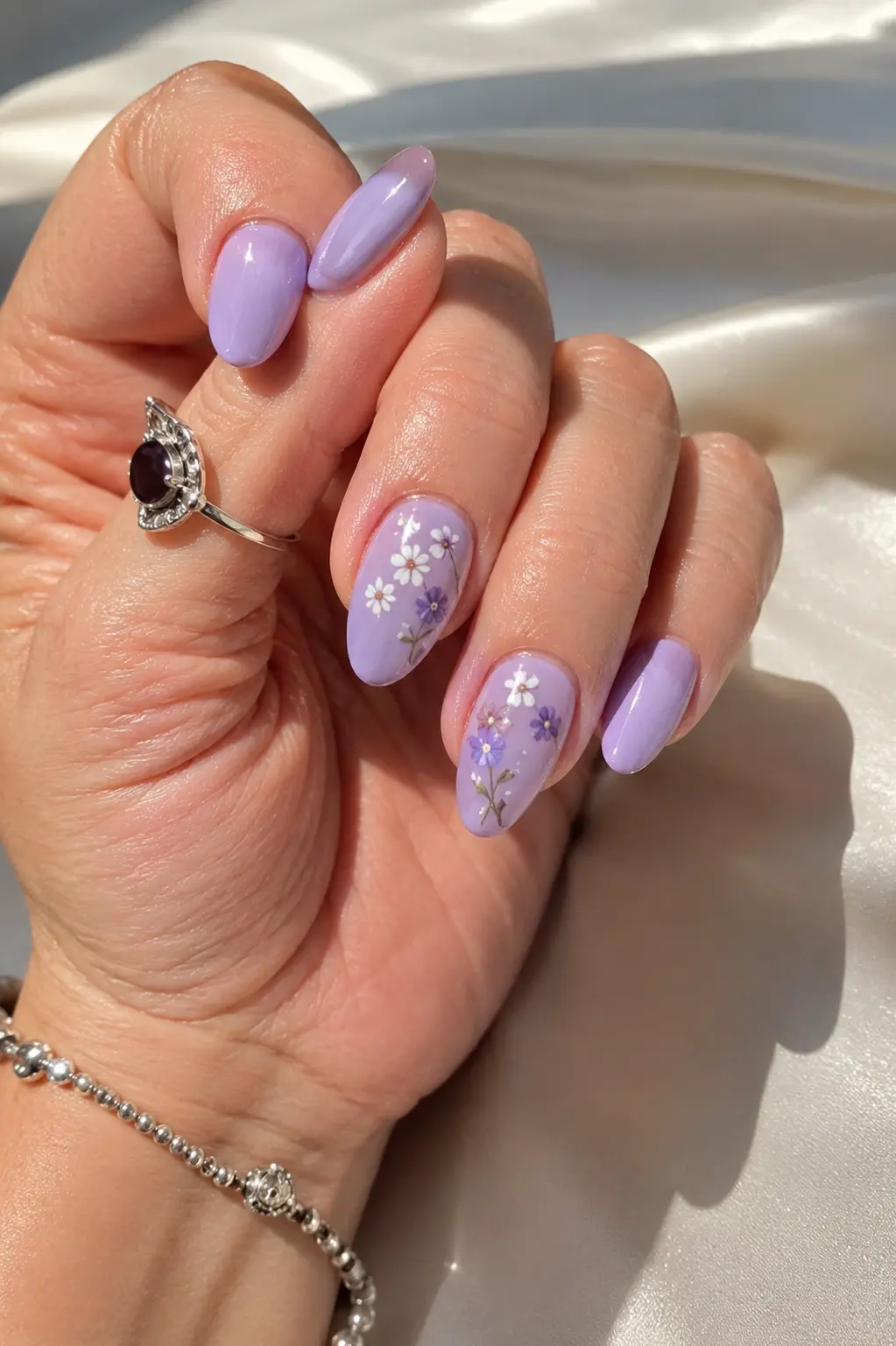 Close-up of glossy pastel lavender almond nails with small floral nail art, silver jewelry, realistic skin texture, and soft natural sunlight on an ivory background.