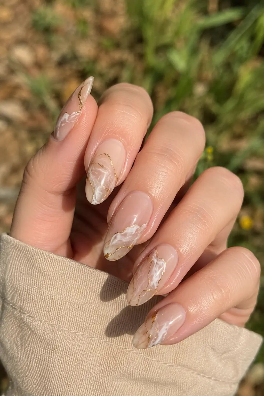 woman's hand with glossy nude almond-shaped nails featuring elegant white and taupe marble nail art accented with delicate gold detailing