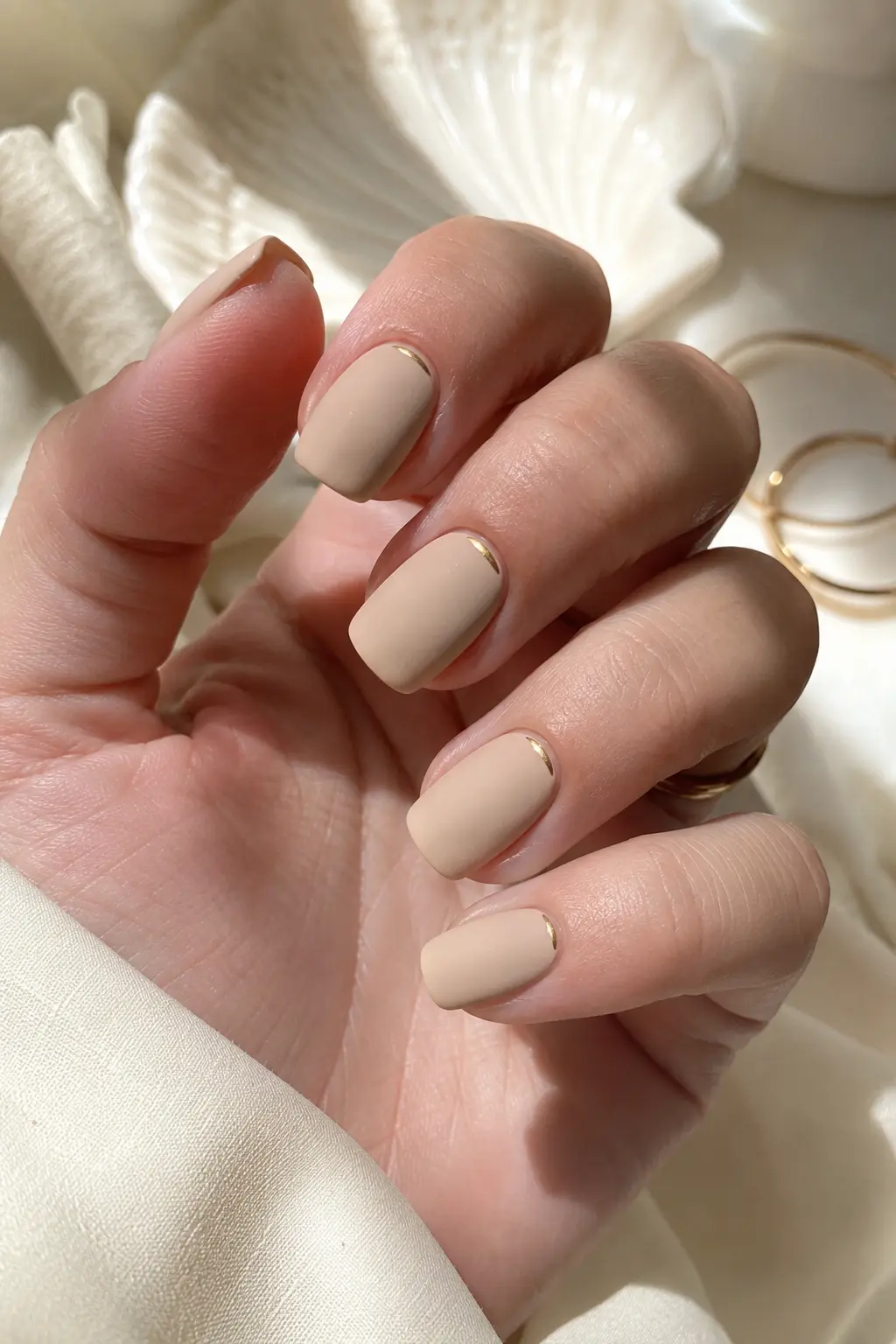 woman's hand with matte beige squoval nails featuring tiny gold accents near the cuticle, photographed in bright natural daylight against an elegant ivory luxury background