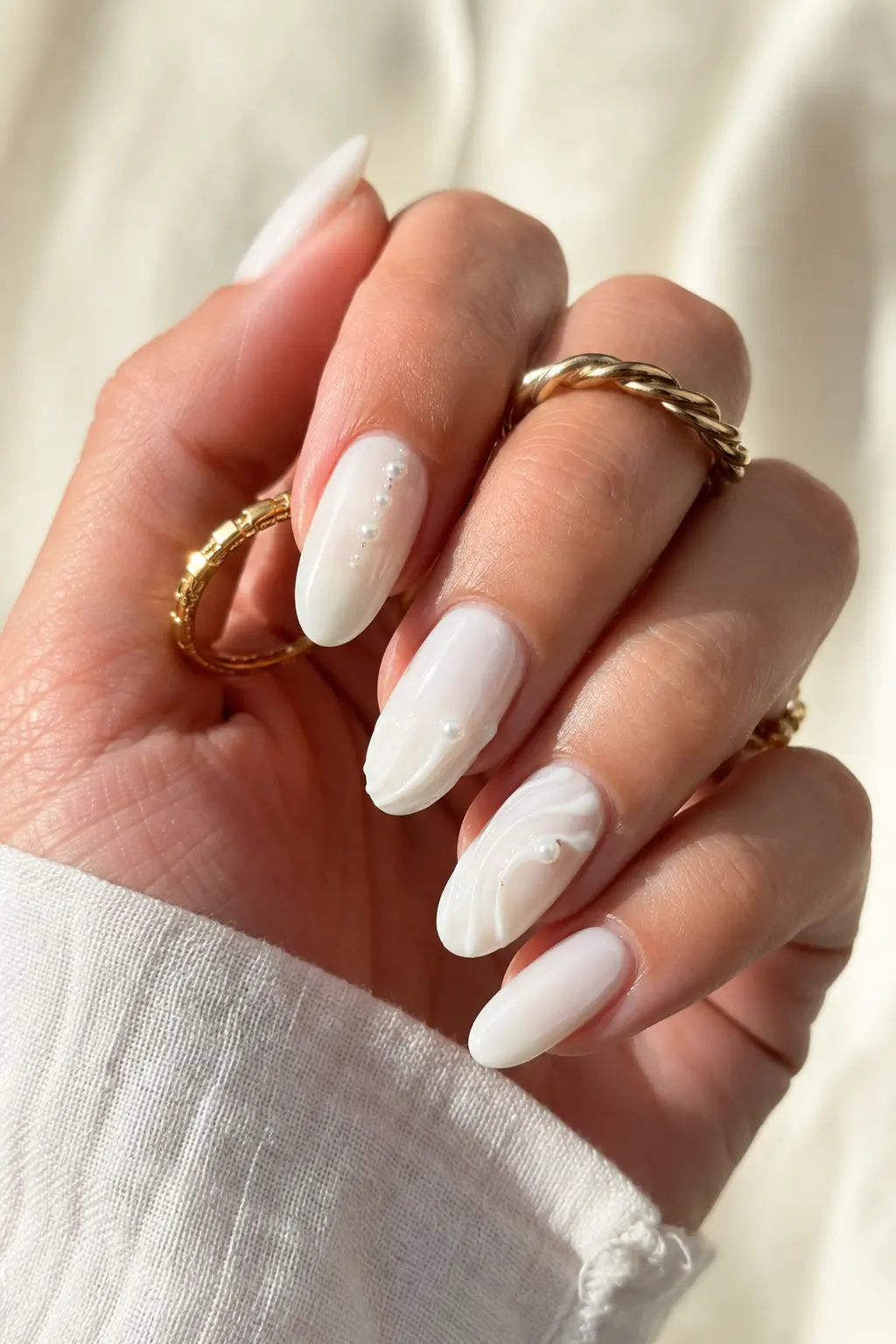 English woman’s hand featuring milky white summer July almond nails with glossy gel polish, pearl nail art details, clean cuticles, gold rings, and a luxury manicure style in soft natural daylight.