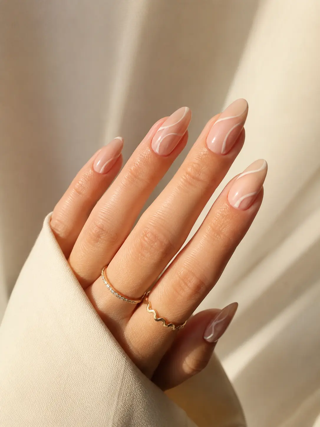 woman’s hand featuring minimal nude summer nails with glossy beige polish, delicate white line art, almond-shaped nails, clean cuticles, gold rings, and soft natural daylight on a cream background.