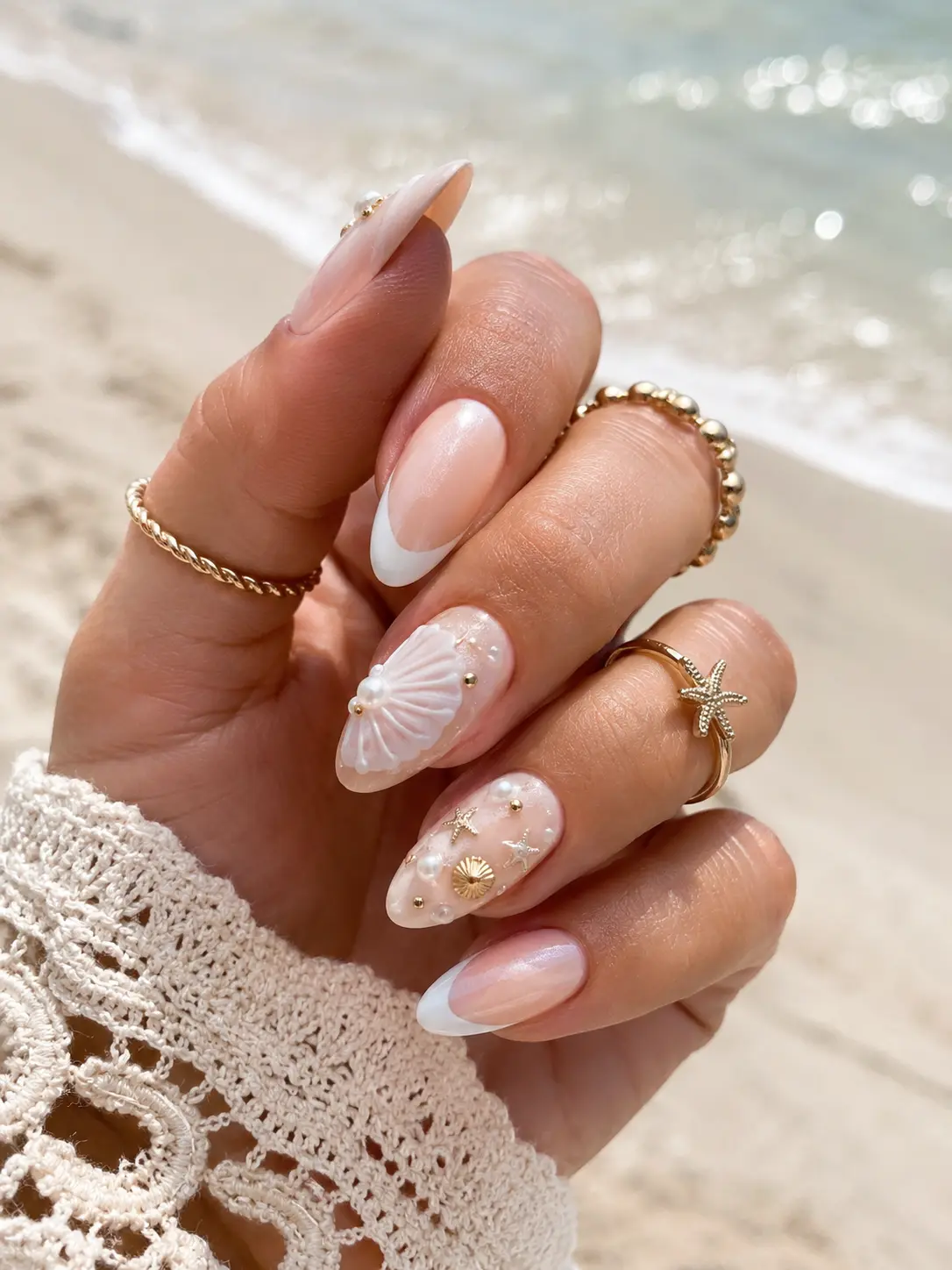 Close-up of a real woman’s hand featuring seashell pearl summer nail designs with glossy nude and white almond nails, pearl accents, tiny seashell nail art, gold rings, and a luxury beach-inspired manicure in natural sunlight.