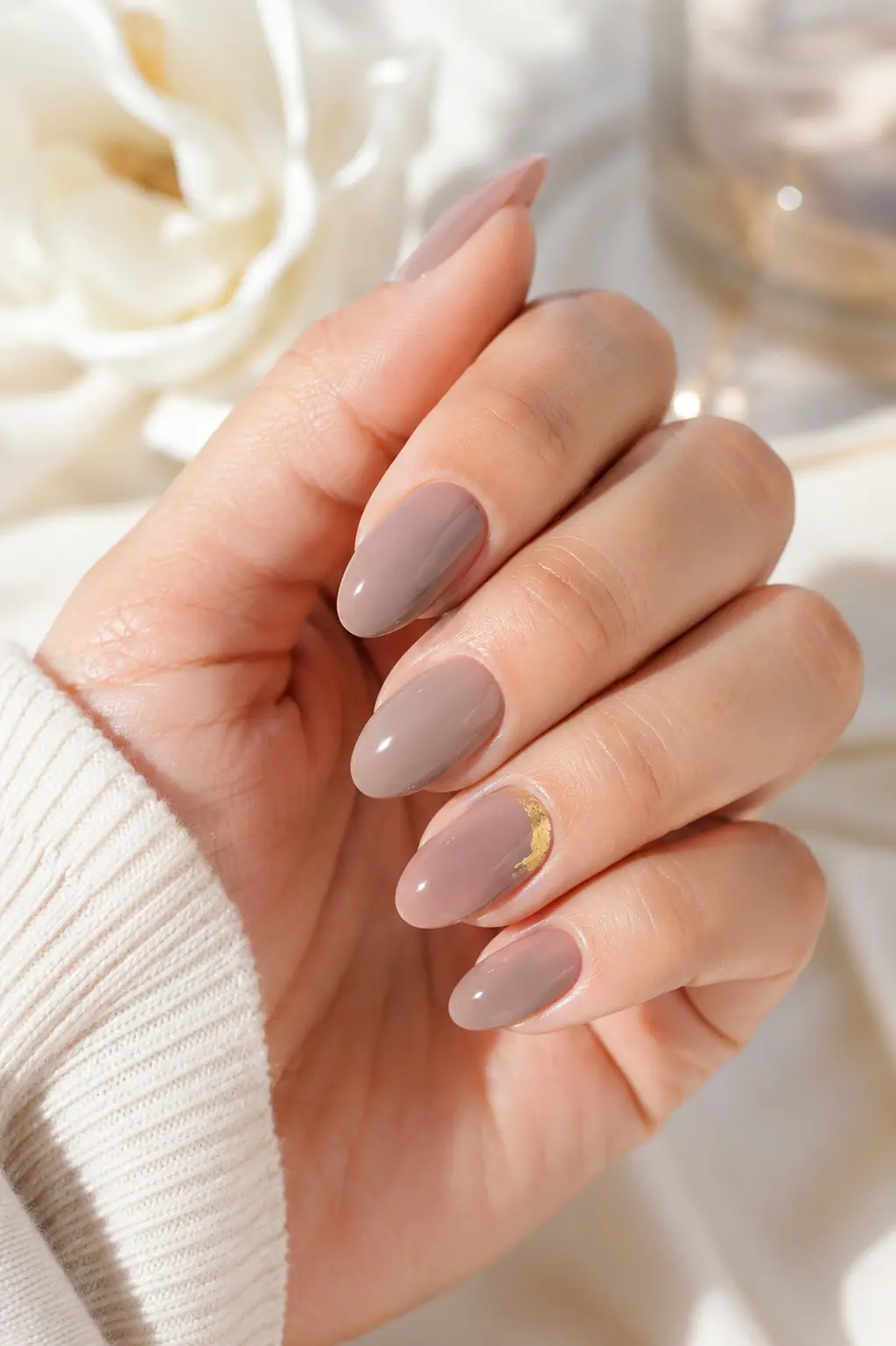 woman's hand with glossy soft taupe almond-shaped nails and a tiny gold foil accent, photographed in bright natural daylight against an elegant ivory and cream luxury background.