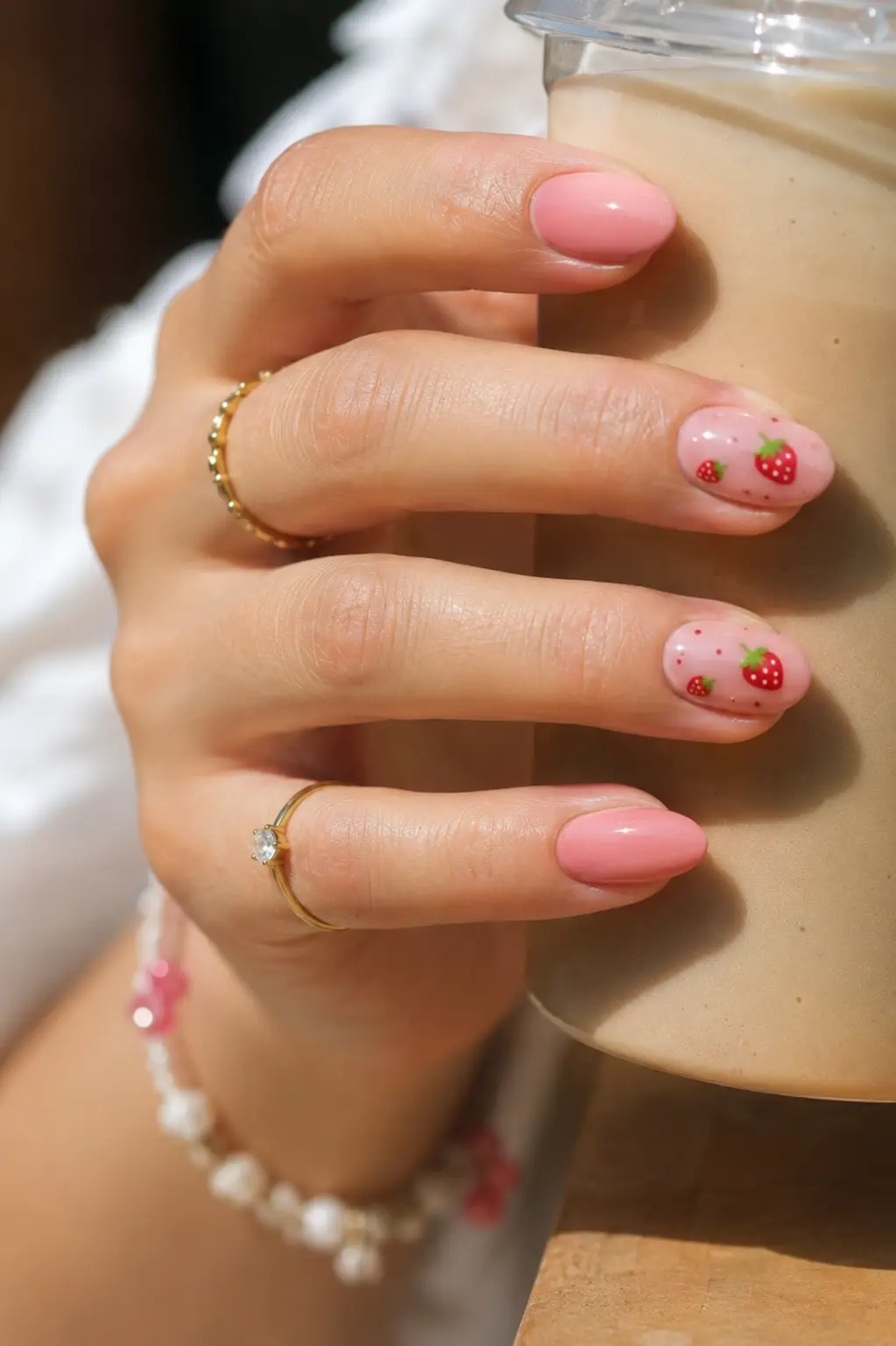 Close-up of a real woman’s hand holding a cup, featuring glossy strawberry pink summer nails with tiny strawberry nail art, almond-shaped gel manicure, gold rings, natural skin texture, and bright daylight.