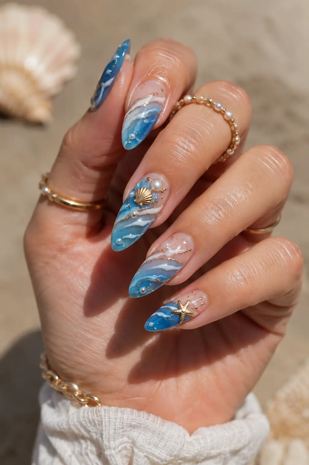 Ocean blue summer July nail design on real women’s hands featuring glossy almond nails, wave nail art, pearl accents, seashell details, gold jewelry, and a beach-inspired manicure look.