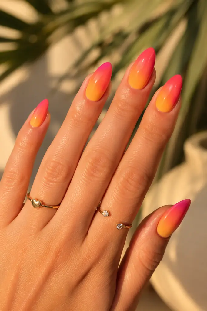 English woman’s hand with glossy sunset ombre summer nails featuring orange, coral, pink, and yellow gradient almond-shaped gel polish, delicate gold jewelry, and golden natural sunlight.